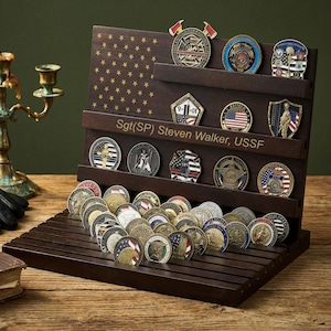 Puede incluir: Un expositor de madera oscura con múltiples estantes, que contiene una colección de monedas militares. El expositor tiene un diseño de bandera estadounidense y el nombre "Sgt(SP) Steven Walker, USSF". También hay un candelabro de latón antiguo, guantes de cuero y un libro.