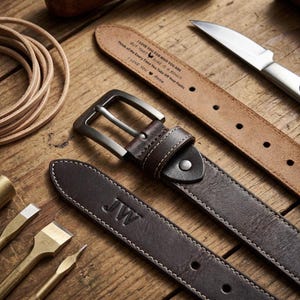 May include: Three leather belts with metal buckles and white stitching are displayed. One belt is embossed with the initials "JW", and another has a personalized message. The belts are brown and tan, with a knife and leatherworking tools in the background.