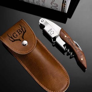 Könnte beinhalten: Ein braunes Lederetui für einen Weinöffner und ein Korkenzieher mit Holzgriff. Der Korkenzieher hat eine silberne Klinge und die Worte "Personalised Text" sind auf dem Griff aufgedruckt. Das Etui hat die Buchstaben "GB" und ein Hirschgeweih-Design.