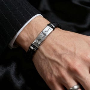 Puede incluir: Pulsera de cuero trenzado negro con una placa plateada grabada con los nombres "Juliet" y "Harry" y un pequeño gráfico. La pulsera se asegura con un cierre plateado y se lleva en la muñeca.