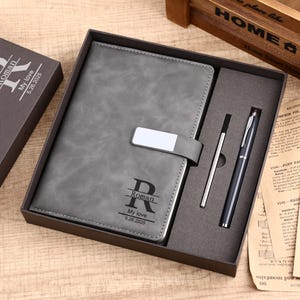 Puede incluir: Un juego de diario y bolígrafo de polipiel gris en una caja de regalo. El diario tiene un cierre plateado y está grabado con el nombre "Roman", las palabras "My love" y la fecha "5.26.2025". El juego incluye un bolígrafo y un lápiz táctil.