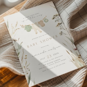 May include: Baby shower invitation with a soft, neutral color palette. The design features a clothesline with baby clothes, floral accents, and the text "Baby Shower" with the honoree's name and event details.