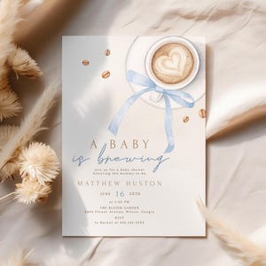 May include: A baby shower invitation with a coffee cup design. The invitation features the text "A baby is brewing" in elegant script, along with event details. The design includes a coffee cup with a heart-shaped latte art and a blue ribbon.