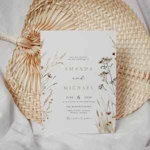 May include: A wedding invitation with floral illustrations and the names Amanda and Michael. The invitation is on a light background with text that reads "June 6 2026" and "The Bloom Garden". The invitation is placed on a woven fan.
