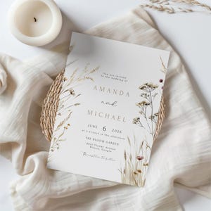 May include: A wedding invitation with the names "AMANDA" and "MICHAEL" in gold lettering, surrounded by floral illustrations. The invitation includes the date "JUNE 6 2026" and the venue "THE BLOOM GARDEN". A white candle and a woven basket are also visible.