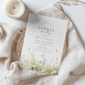 May include: A white invitation card with the text "GARDEN party" and "35th BIRTHDAY" in black. The card features a floral design at the bottom and is placed on a beige cloth with a woven basket and a white candle.