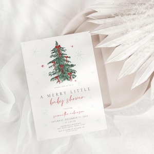 May include: A baby shower invitation with a watercolor Christmas tree design. The invitation reads "A Merry Little Baby Shower" in red script, with event details below. The card is on a white plate with white fabric and decorative elements.