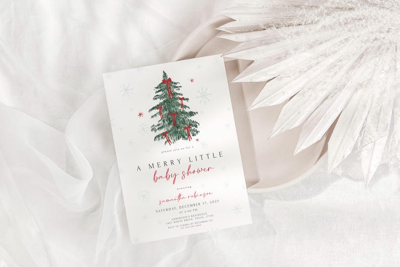 May include: A baby shower invitation with a watercolor Christmas tree design. The invitation reads "A Merry Little Baby Shower" in red script, with event details below. The card is on a white plate with white fabric and decorative elements.