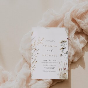 May include: A wedding invitation with the names Amanda and Michael, and the date June 6, 2026. The invitation is decorated with floral elements and rests on a soft, draped fabric.