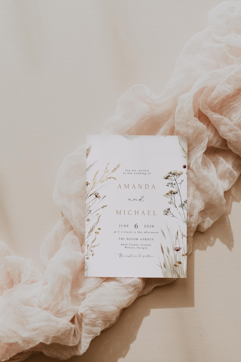 May include: A wedding invitation with the names Amanda and Michael, and the date June 6, 2026. The invitation is decorated with floral elements and rests on a soft, draped fabric.
