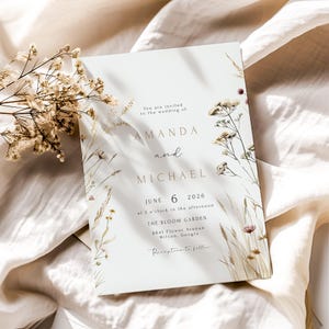 May include: A wedding invitation with the names "AMANDA" and "MICHAEL" in elegant gold lettering. The invitation includes the date "JUNE 6 2026" and venue details, framed by delicate floral illustrations in neutral tones. The text "Reception to follow" is also included.