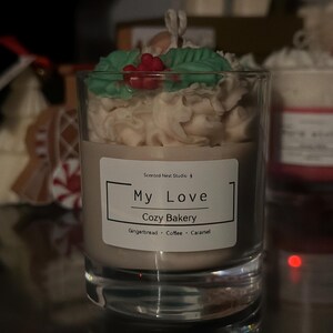 Pu&ograve; includere: Una candela in un bicchiere trasparente, sormontata da un design cremoso, foglie verdi e bacche rosse. L'etichetta dice "My Love Cozy Bakery" con profumi di pan di zenzero, caff&egrave; e caramello. La candela &egrave; in un contenitore di vetro.