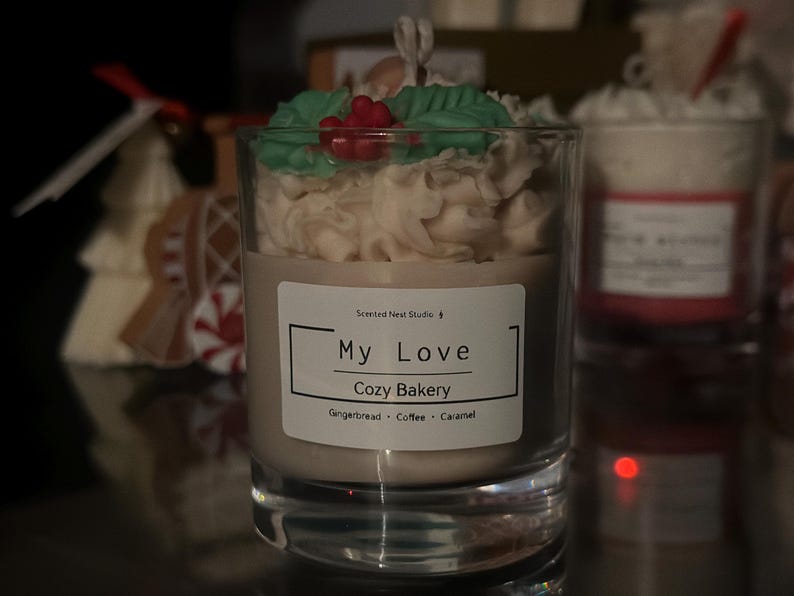 Pu&ograve; includere: Una candela in un bicchiere trasparente, sormontata da un design cremoso, foglie verdi e bacche rosse. L'etichetta dice "My Love Cozy Bakery" con profumi di pan di zenzero, caff&egrave; e caramello. La candela &egrave; in un contenitore di vetro.