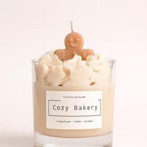 Pu&ograve; includere: Una candela profumata in cera di soia in un bicchiere trasparente, etichettata "Cozy Bakery". La candela presenta una base di cera marrone chiaro, sormontata da accenti di cera bianca e una piccola figura di omino di pan di zenzero. L'etichetta elenca pan di zenzero, caff&egrave; e caramello.