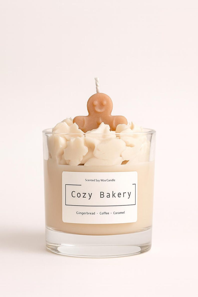 Pu&ograve; includere: Una candela profumata in cera di soia in un bicchiere trasparente, etichettata "Cozy Bakery". La candela presenta una base di cera marrone chiaro, sormontata da accenti di cera bianca e una piccola figura di omino di pan di zenzero. L'etichetta elenca pan di zenzero, caff&egrave; e caramello.