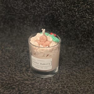 Pu&ograve; includere: Una candela in vetro con un design a omino di pan di zenzero. La candela ha una base di cera marrone chiaro ed &egrave; sormontata da elementi decorativi bianchi, verdi e rossi. L'etichetta recita "Cozy Bakery".