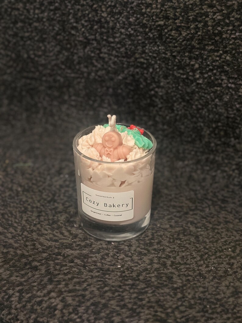 Pu&ograve; includere: Una candela in vetro con un design a omino di pan di zenzero. La candela ha una base di cera marrone chiaro ed &egrave; sormontata da elementi decorativi bianchi, verdi e rossi. L'etichetta recita "Cozy Bakery".