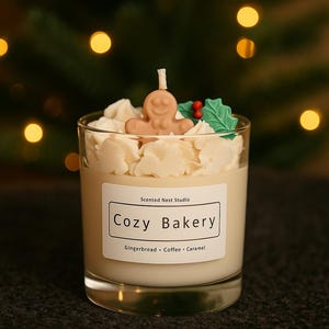 Pu&ograve; includere: Candela profumata in un bicchiere trasparente, sormontata da un omino di pan di zenzero. L'etichetta recita "Cozy Bakery" con "Gingerbread, Coffee, Caramel" sotto. Decorata con glassa bianca e agrifoglio.