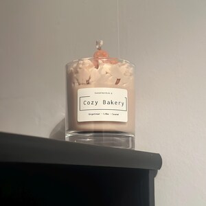 Pu&ograve; includere: Una candela profumata in un barattolo di vetro trasparente, sormontata da cera bianca e una piccola decorazione rotonda color arancione. L'etichetta recita "Cozy Bakery" con note olfattive di Pan di zenzero, Caff&egrave; e Caramello. La candela &egrave; su una superficie nera.