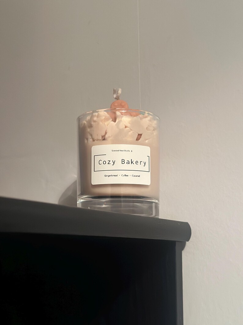 Pu&ograve; includere: Una candela profumata in un barattolo di vetro trasparente, sormontata da cera bianca e una piccola decorazione rotonda color arancione. L'etichetta recita "Cozy Bakery" con note olfattive di Pan di zenzero, Caff&egrave; e Caramello. La candela &egrave; su una superficie nera.