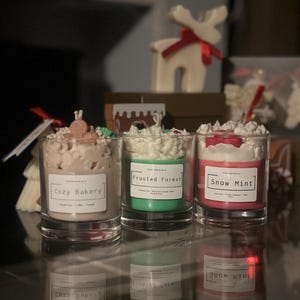 Puede incluir: Tres velas perfumadas en frascos de vidrio con coberturas de cera batida. Las etiquetas dicen "Cozy Bakery", "Frosted Forest" y "Snow Mint". Una decoración de reno blanco con un lazo rojo está en el fondo.