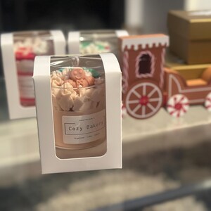 Pu&ograve; includere: Una candela in un barattolo di vetro con l'etichetta "Cozy Bakery" &egrave; presentata in una scatola bianca. La candela &egrave; decorata con glassa color crema e piccoli accenti arancioni e verdi. Un trenino di pan di zenzero &egrave; sullo sfondo.