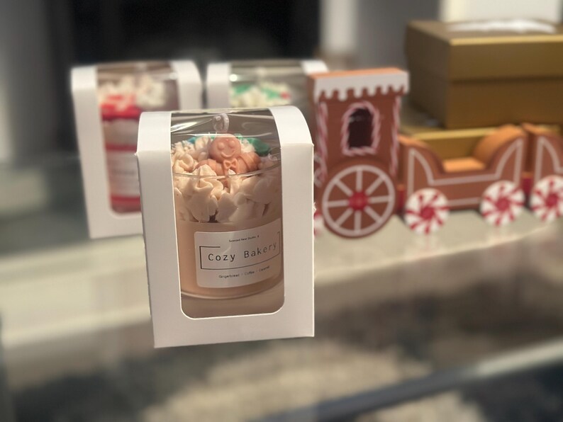 Pu&ograve; includere: Una candela in un barattolo di vetro con l'etichetta "Cozy Bakery" &egrave; presentata in una scatola bianca. La candela &egrave; decorata con glassa color crema e piccoli accenti arancioni e verdi. Un trenino di pan di zenzero &egrave; sullo sfondo.
