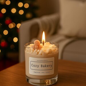 Pu&ograve; includere: Una candela accesa in un barattolo di vetro trasparente, sormontata da un omino di pan di zenzero. L'etichetta recita "Cozy Bakery" con "Gingerbread - Coffee - Caramel" sotto. Lo sfondo &egrave; sfocato, con un albero di Natale e un divano visibili.