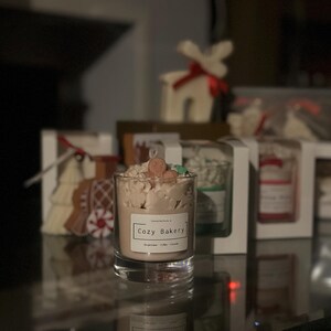 Pu&ograve; includere: Una candela in un barattolo di vetro trasparente con l'etichetta "Cozy Bakery". La candela ha cera marrone chiaro con guarnizioni decorative. Altre confezioni regalo con candele e oggetti decorativi sono sullo sfondo.