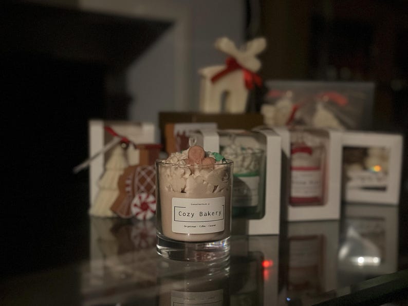 Pu&ograve; includere: Una candela in un barattolo di vetro trasparente con l'etichetta "Cozy Bakery". La candela ha cera marrone chiaro con guarnizioni decorative. Altre confezioni regalo con candele e oggetti decorativi sono sullo sfondo.