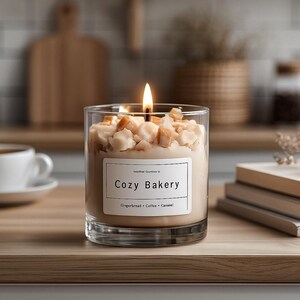 Pu&ograve; includere: Una candela accesa in un barattolo di vetro trasparente con l'etichetta "Cozy Bakery" con note di profumo di pan di zenzero, caff&egrave; e caramello. La candela &egrave; sormontata da pezzi color caramello. Una tazza e un piattino bianchi sono sullo sfondo.