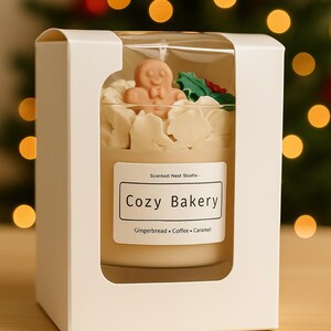 Pu&ograve; includere: Candela profumata in un barattolo di vetro, presentata in una scatola bianca. La candela presenta un omino di pan di zenzero e accenti di agrifoglio. L'etichetta recita "Cozy Bakery" con "Gingerbread, Coffee, Caramel" sotto.