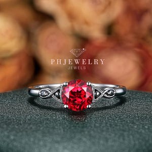 May include: A silver ring with a large, round, bright red gemstone as the centerpiece. The ring features a delicate design with small, clear accent stones on either side of the main stone. The ring is set against a blurred background with the text "PHJEWELRY JEWELS".