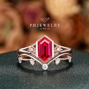 May include: Rose gold ring set featuring a large, rectangular, red gemstone as the centerpiece. The ring is accented with smaller, clear gemstones and floral details. The brand name "PHJEWELRY JEWELS" is visible in the background.