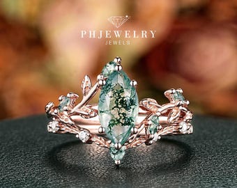 May include: A rose gold ring with a marquise-cut moss agate gemstone. The ring features a nature-inspired design with leaf and vine details, and small accent stones. The ring is set against a dark green surface.