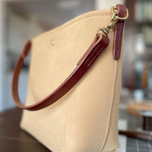 May include: A beige leather handbag with a burgundy strap and accents. The bag has a rectangular shape with a rounded bottom and is secured with a brass clasp. The strap is stitched and attached with a swivel hook.