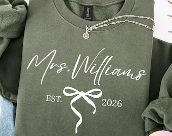 Può includere: Una felpa verde oliva con la scritta "Mrs. Williams EST. 2026" in corsivo bianco. Una collana d'argento con un ciondolo è drappeggiata sulla felpa. La felpa è piegata, con una porzione di denim blu visibile.