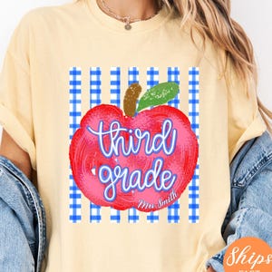 May include: A butter yellow t-shirt featuring a graphic of a red apple with a green leaf and brown stem. The apple is layered over a blue and white gingham striped background. The text "third grade" is written in blue, with "Mrs. Smith" below.