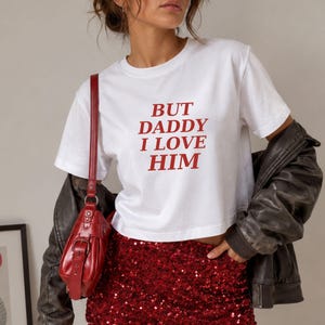 May include: A white short-sleeved t-shirt with the red text "BUT DADDY I LOVE HIM" printed on the front, paired with a red sequined mini skirt, a dark grey leather jacket, and a red shoulder bag with a buckle strap.