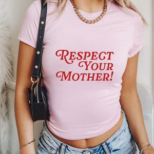 May include: A light pink t-shirt with the text "Respect Your Mother!" in red script. The shirt has short sleeves and is paired with blue jeans. A black purse with a gold chain is visible.