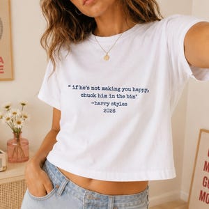 May include: A white short-sleeved t-shirt featuring dark blue text that reads: "if he's not making you happy, chuck him in the bin" -harry styles 2026. The shirt is paired with light blue denim shorts and a gold chain necklace.
