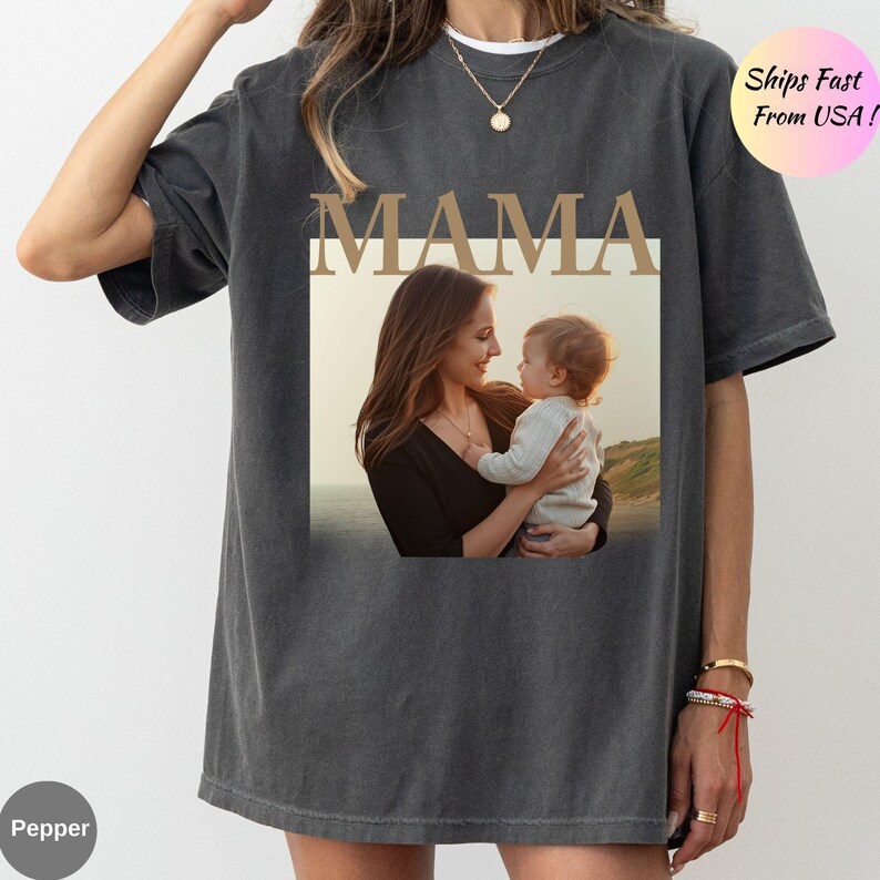 May include: Dark gray t-shirt with the word "MAMA" in gold lettering above a framed photo of a mother holding a baby. The photo is set against a blurred outdoor background. The shirt is a relaxed fit.
