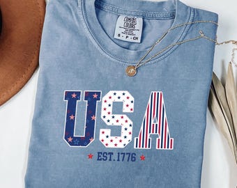 May include: A light blue Comfort Colors t-shirt with the word "USA" in a patriotic design. The "U" and "A" are decorated with red, white, and blue stars and stripes. The text "EST. 1776" is below the word "USA". A gold necklace is draped across the shirt.