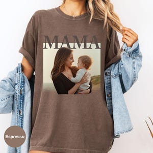 May include: Espresso-colored t-shirt featuring the word "MAMA" above a framed photograph of a mother and child. The photograph shows a mother holding a baby, with a beach scene in the background. The shirt has rolled-up sleeves.