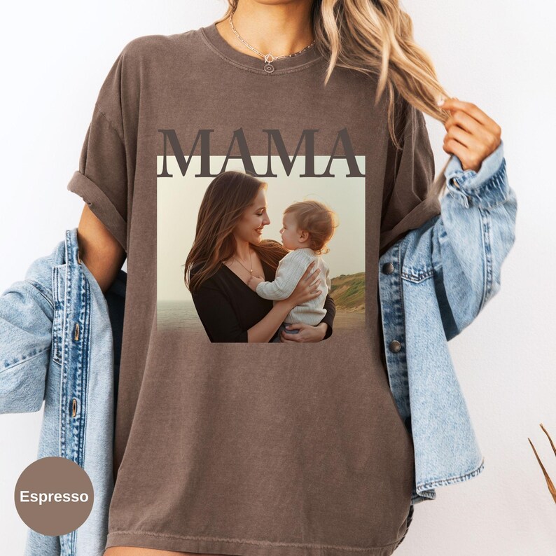 May include: Espresso-colored t-shirt featuring the word "MAMA" above a framed photograph of a mother and child. The photograph shows a mother holding a baby, with a beach scene in the background. The shirt has rolled-up sleeves.