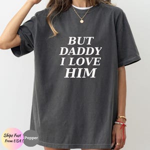 May include: A charcoal grey short-sleeve t-shirt featuring the white text BUT DADDY I LOVE HIM printed in a bold, serif font across the chest. The garment has a crew neckline and a relaxed, oversized fit with a slightly faded, vintage-style texture.