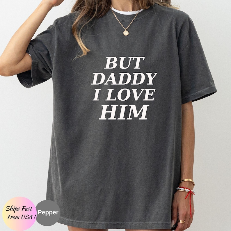 May include: A charcoal grey short-sleeve t-shirt featuring the white text BUT DADDY I LOVE HIM printed in a bold, serif font across the chest. The garment has a crew neckline and a relaxed, oversized fit with a slightly faded, vintage-style texture.
