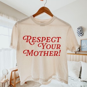 Peut inclure: T-shirt crème avec l'inscription "Respect Your Mother!" en rouge, en gras et en cursive. Le t-shirt est suspendu à un cintre en bois. Le t-shirt a des manches courtes et un col rond.