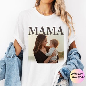 May include: White t-shirt with the word "MAMA" in a bold, serif font. Below the word is a photograph of a mother holding a baby. The shirt has rolled-up sleeves and is paired with a denim jacket.