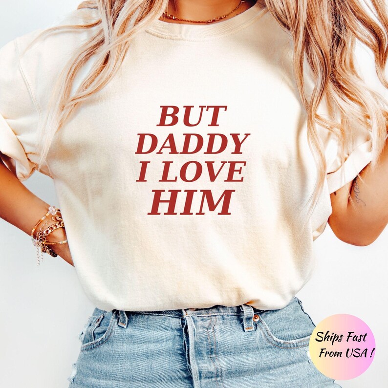 May include: A cream-colored short-sleeve t-shirt featuring the text BUT DADDY I LOVE HIM printed in a dark red serif font across the chest. The shirt is paired with blue denim jeans and worn with a gold chain necklace and gold bracelets.